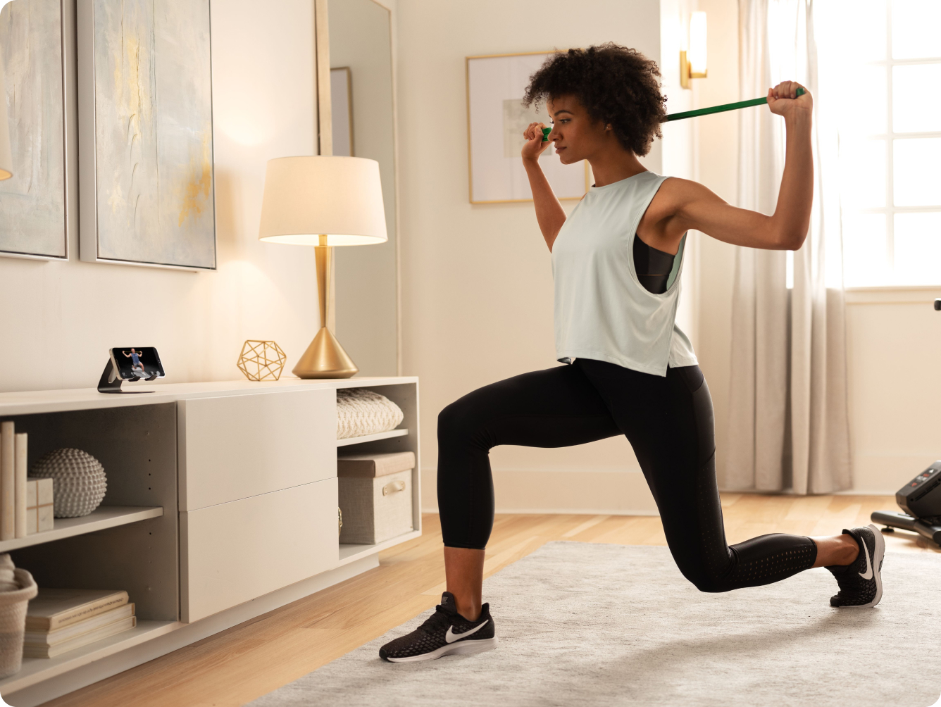 Black woman exercising in her living room with a bar behind her head watching the iFIT app on a smart phone.